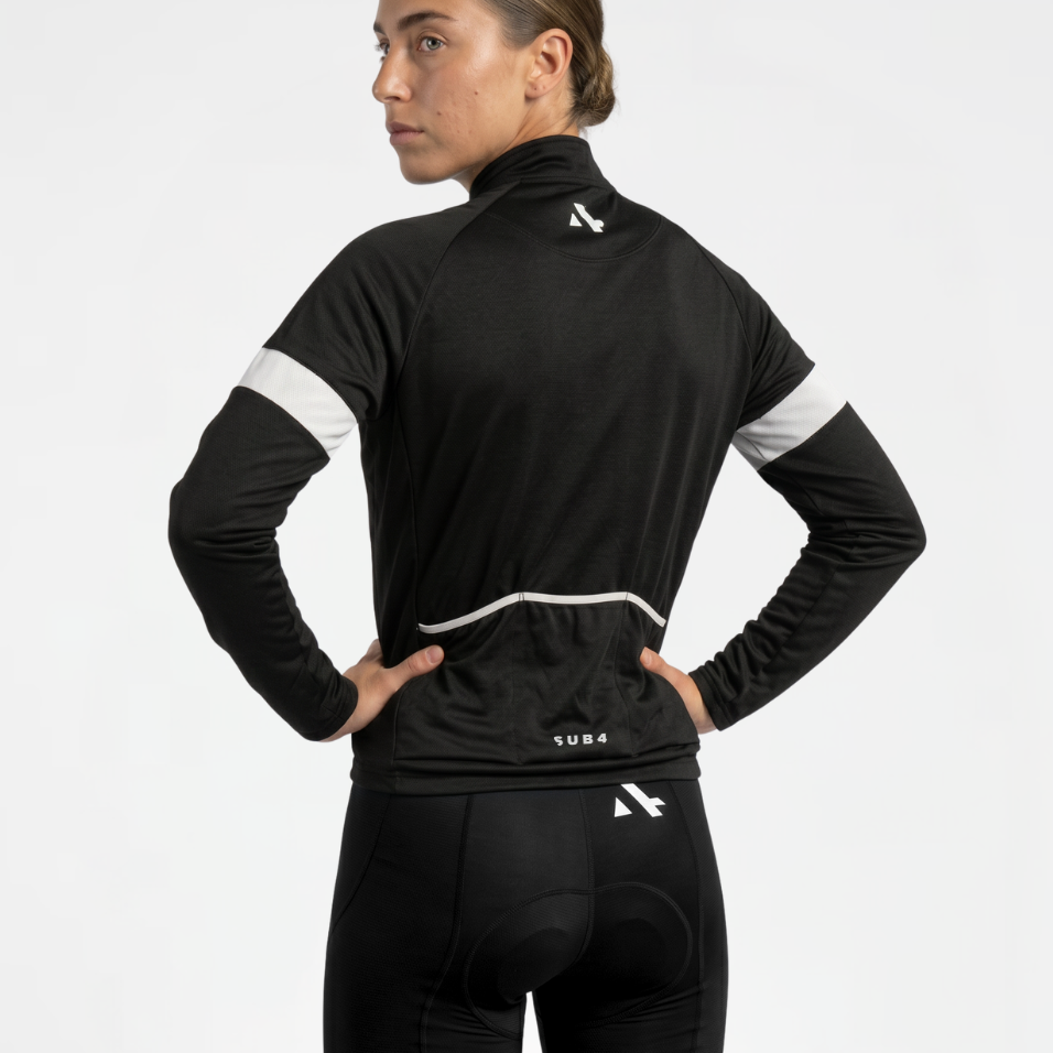 Back view of woman wearing a black cycling thermal jacket and black cycling shorts with white reflective accents on a light gray background
