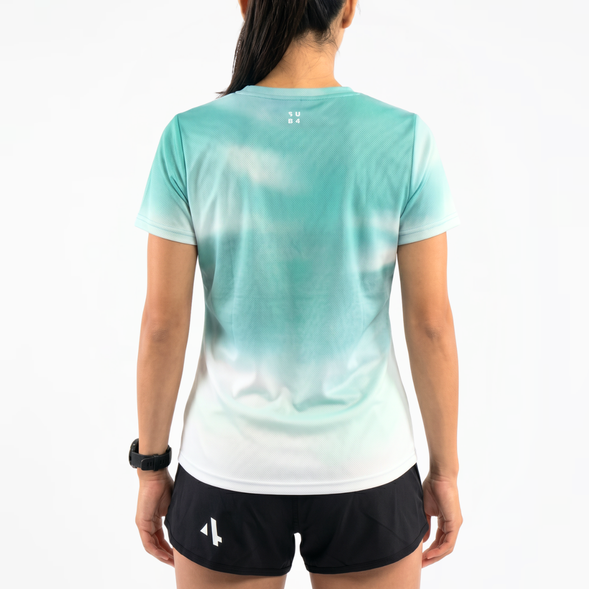 Back view of woman wearing a gradient teal running t-shirt and black running shorts on a white background