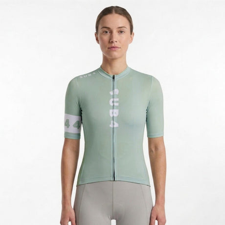 A woman modeling a light olive sage green short-sleeve cycling jersey with vertical white 'SUB4' branding on the front and a white armband detail, paired with grey cycling shorts against a plain white background.