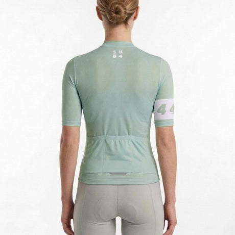 A woman viewed from the back wearing a light olive sage green short-sleeve cycling jersey and grey cycling shorts against a plain white background.