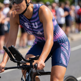 A female athlete riding a triathlon bike outdoors, wearing a sleeveless one-piece suit with a pink, purple, and navy geometric pattern and the letters 'SUB4' across the chest.
